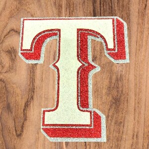May include: A white letter T with a red outline and glitter. The letter is on a wood background.