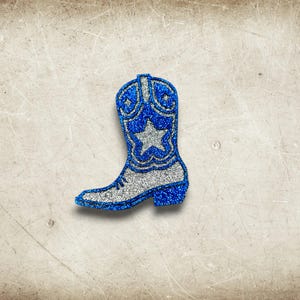 May include: A glittery blue and silver cowboy boot-shaped decoration. The boot features a silver star and decorative patterns on the upper shaft. The boot's outline and details are accented with sparkling blue glitter, creating a festive and eye-catching design.
