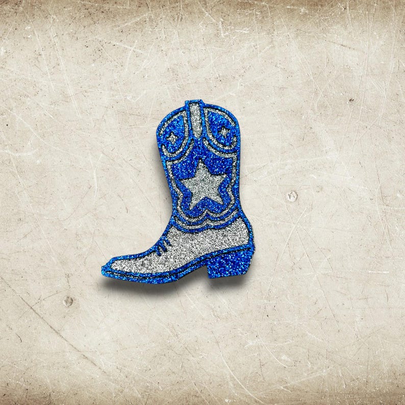 May include: A glittery blue and silver cowboy boot-shaped decoration. The boot features a silver star and decorative patterns on the upper shaft. The boot's outline and details are accented with sparkling blue glitter, creating a festive and eye-catching design.