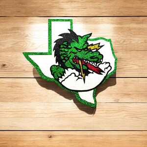 May include: Green glitter outline of the state of Texas with a green, yellow, and red cartoon dragon inside. The dragon is breathing fire and has a menacing expression.