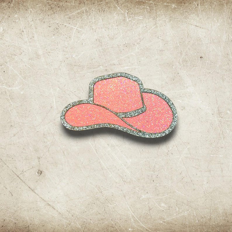 May include: A pink glitter cowboy hat with a silver glitter trim. The hat is a decorative item, possibly a brooch or embellishment, and is set against a neutral background.