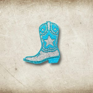 May include: A glittery turquoise cowboy boot with silver accents and a star design. The boot has a classic western silhouette, with a pointed toe and a small heel. The boot is a decorative item.