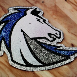 May include: A silver and blue glitter iron-on patch featuring a white horse head with a blue mane. The horse is facing the right side of the image.