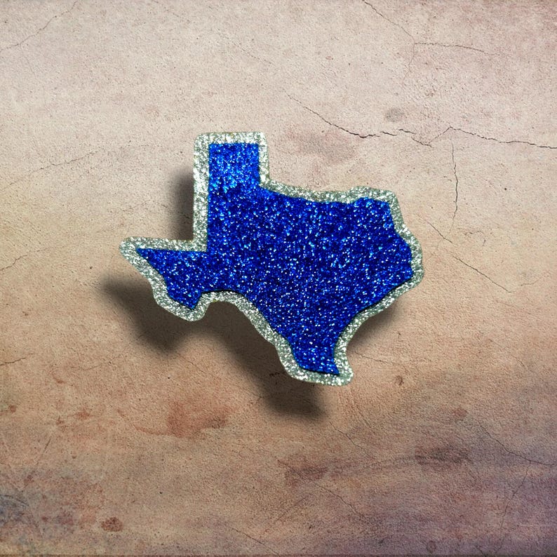 May include: A blue glitter Texas-shaped ornament with a silver glitter outline. The ornament is set against a textured, light brown background. The Texas-shaped item is a decorative item.
