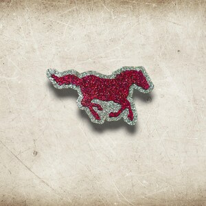 May include: A red glitter horse-shaped embellishment with a silver outline. The horse is depicted in a running pose, with its legs extended. The embellishment is on a textured, off-white background.