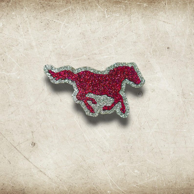 May include: A red glitter horse-shaped embellishment with a silver outline. The horse is depicted in a running pose, with its legs extended. The embellishment is on a textured, off-white background.