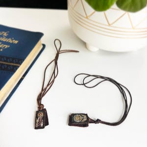 May include: Two small, rectangular religious pendants with brown cords rest on a white surface. The pendants feature detailed artwork and are attached to braided cords. A blue book and a white pot with a gold geometric design are in the background.