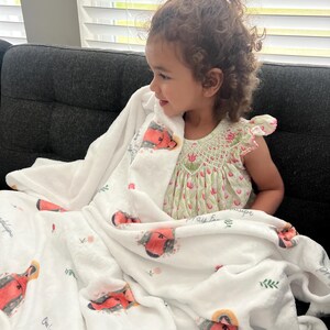 May include: A white, plush blanket with a red and orange illustration of Our Lady of Guadalupe, and the text "Our Lady of Guadalupe, pray for us." The blanket has a floral design and is wrapped around a child.