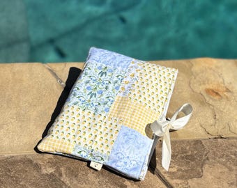 Peut inclure: Une pochette en tissu rectangulaire présentant un motif patchwork composé de fleurs bleu clair, de citrons jaunes et de carreaux vichy jaunes. La pochette est fermée par un ruban blanc noué en nœud et comporte une petite étiquette en tissu blanc.