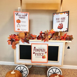 May include: A white wooden cart with black wheels and a chalkboard sign that says "Grown Locally Pumpkin Patch Serving Treats and Hot Drinks". There are pumpkins, autumn leaves, and signs that say "Pick Your Pumpkin!" and "Pay Here Cash Only!"