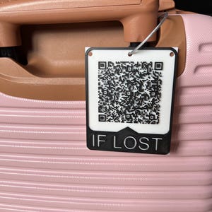 Könnte beinhalten: Nahaufnahme eines rosa Koffers mit braunem Griff. Ein weißes Etikett mit einem QR-Code und dem Text "IF LOST" ist am Griff befestigt. Der Koffer hat eine gerippte Textur.