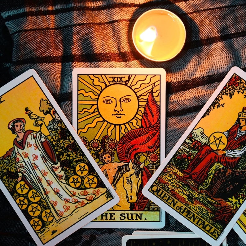 May include: Three tarot cards, The Sun, Queen of Pentacles, and The World, laid out on a patterned fabric with a lit candle in the background. The Sun card features a bright yellow sun with a face, surrounded by rays of light. The Queen of Pentacles card shows a queen holding a pentacles coin, sitting on a throne with a red robe. The World card depicts a woman standing in a circle with a wand in her hand.