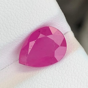 May include: A faceted, pear-shaped ruby gemstone. The gem is a vibrant pinkish-red color and sits on a white surface. The cut of the stone reveals multiple facets, enhancing its brilliance. The gemstone is a focal point, showcasing its clarity and color.