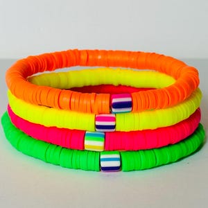 May include: A stack of four stretch bracelets made of neon-colored polymer clay disc beads. The bracelets feature vibrant orange, yellow, pink, and green colors, each accented with a single square bead displaying horizontal stripes in various colors.
