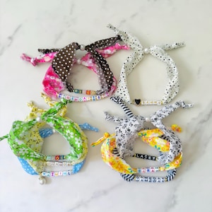 May include: Four fabric bracelets with personalized beaded names and phrases. The bracelets feature various patterns, including polka dots, floral prints, and stripes, in colors like pink, white, green, yellow, and black. Each bracelet has a decorative knot.