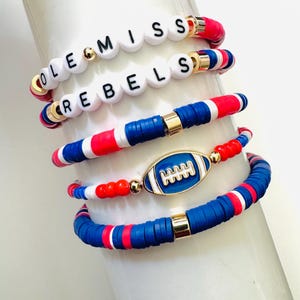 Puede incluir: Una pila de pulseras en rojo, blanco y azul. Dos pulseras deletrean "OLE MISS REBELS" con cuentas de letras blancas. Otras pulseras presentan un dije de fútbol y patrones de colores alternos. Se ven detalles dorados.