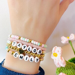 Op de afbeelding: Een stapel armbanden met kralen in pastelkleuren en gouden accenten. Eén armband spelt "TAYLOR" met witte letterkralen en een gouden hartbedel. Een andere armband spelt "EMILY". De armbanden zitten om een pols.