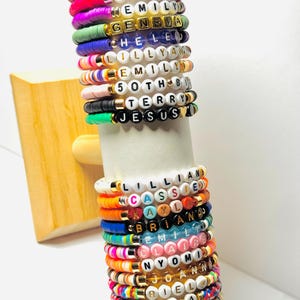 May include: A collection of colorful beaded bracelets with names and phrases spelled out in white letter beads. The bracelets are stacked on a white cylindrical display. The names include GIANA, EMILY, GENEVA, CHELED, LILLYAN, TERRY, and JESUS.