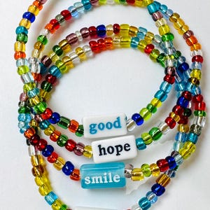 May include: Colorful beaded bracelets with the words "good", "hope", "smile", and "sweet". The bracelets are made of small, multi-colored beads in shades of red, yellow, blue, green, and purple. The words are on white and blue rectangular beads.