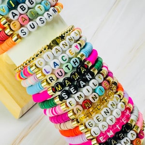 Puede incluir: Una colección de pulseras de cuentas coloridas con nombres escritos con cuentas de letras blancas. Las pulseras presentan detalles dorados y una variedad de colores, incluyendo rosa, verde, azul y naranja. Las pulseras están apiladas.