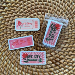 May include: A collection of rectangular paper tickets with pink and red designs. One ticket reads Date Night for two, another Love Day Feb 14, and others Love City Admit One $5. Some tickets are loose, and a stack is inside a clear plastic sleeve.