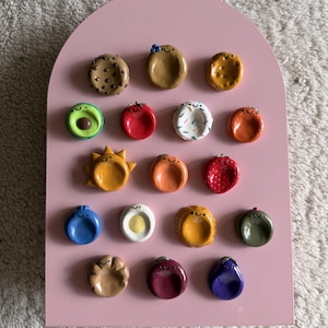 May include: A pink arched display board featuring seventeen small, circular, hand-sculpted clay charms. Each charm depicts a unique food item or object, including a cookie, avocado, sun, egg, and various fruits, all finished with a glossy glaze and simple, hand-painted smiling faces.