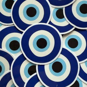 May include: A pile of circular stickers with a blue and white evil eye motif. Each sticker has a dark blue border, a white inner ring, a light blue ring, and a black centre. The stickers are overlapping, creating a layered effect.