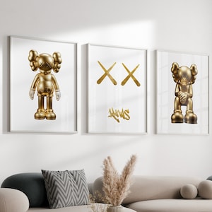 May include: Three framed prints on a white wall. The left and right prints feature a gold figurine of a cartoon character with large eyes and arms crossed. The middle print features a gold graphic of two crossed Xs with the word "NEWS" written in gold below.