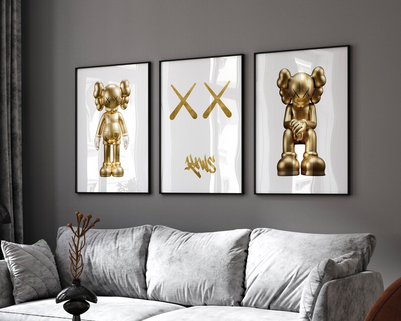 May include: Three framed prints with a white background. The centre print features a gold 'XX' design. The prints on the left and right feature a gold figurine of a character with a blank expression. The figurine on the left is standing and the figurine on the right is sitting.