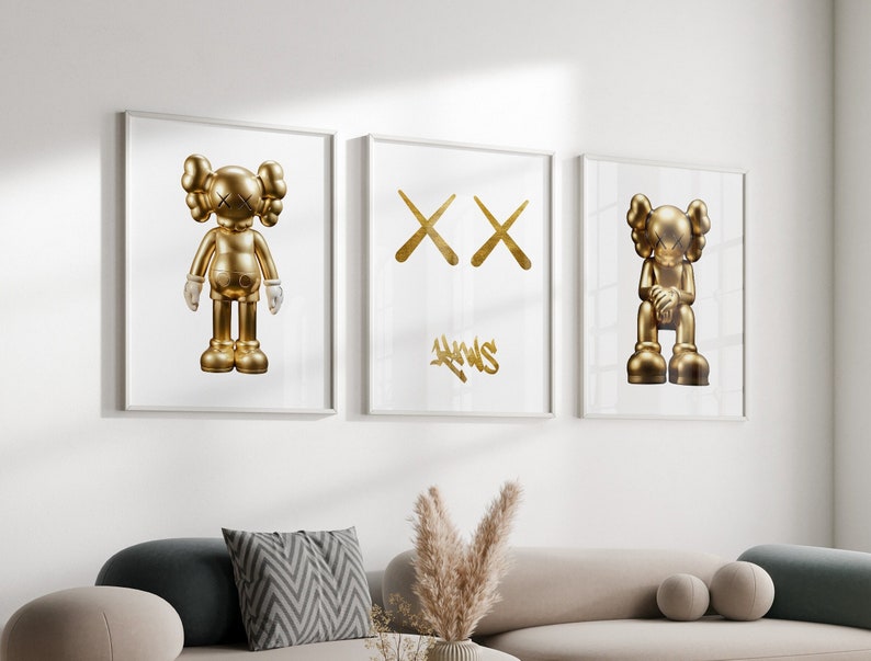 May include: Three framed prints on a white wall. The left and right prints feature a gold figurine of a cartoon character with large eyes and arms crossed. The middle print features a gold graphic of two crossed Xs with the word "NEWS" written in gold below.