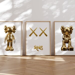 May include: Three framed prints with gold-coloured illustrations. The left and right prints feature a gold-coloured cartoon character with a simple design. The centre print features two gold-coloured X's and the word "KAWS" in gold-coloured graffiti lettering.