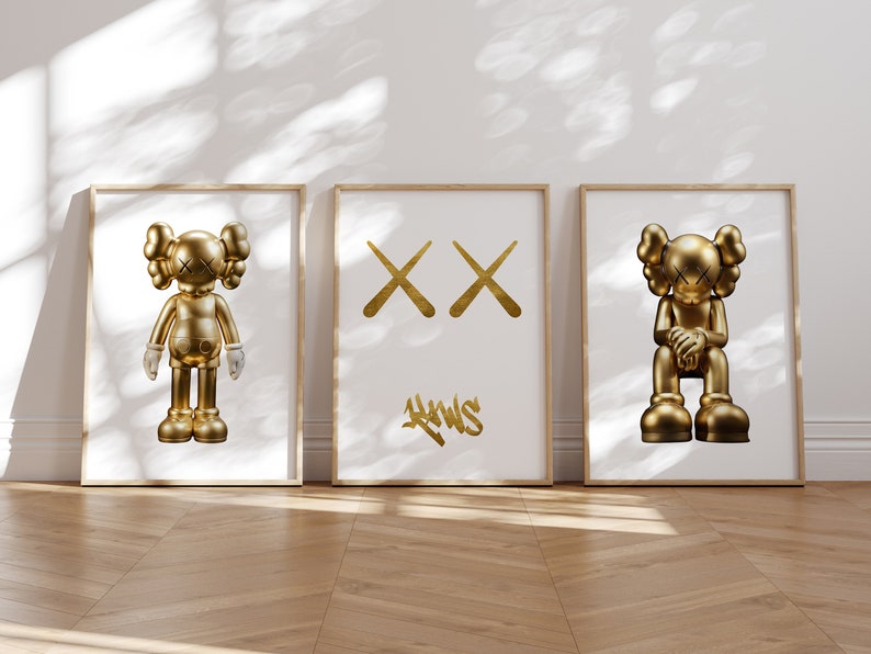 May include: Three framed prints with gold-coloured illustrations. The left and right prints feature a gold-coloured cartoon character with a simple design. The centre print features two gold-coloured X's and the word "KAWS" in gold-coloured graffiti lettering.