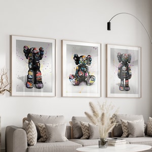May include: Three framed prints of black and white graffiti art featuring a cartoon character. The character is depicted in three different poses: standing, sitting, and with arms raised. The background of each print is a light grey with colourful paint splatters.
