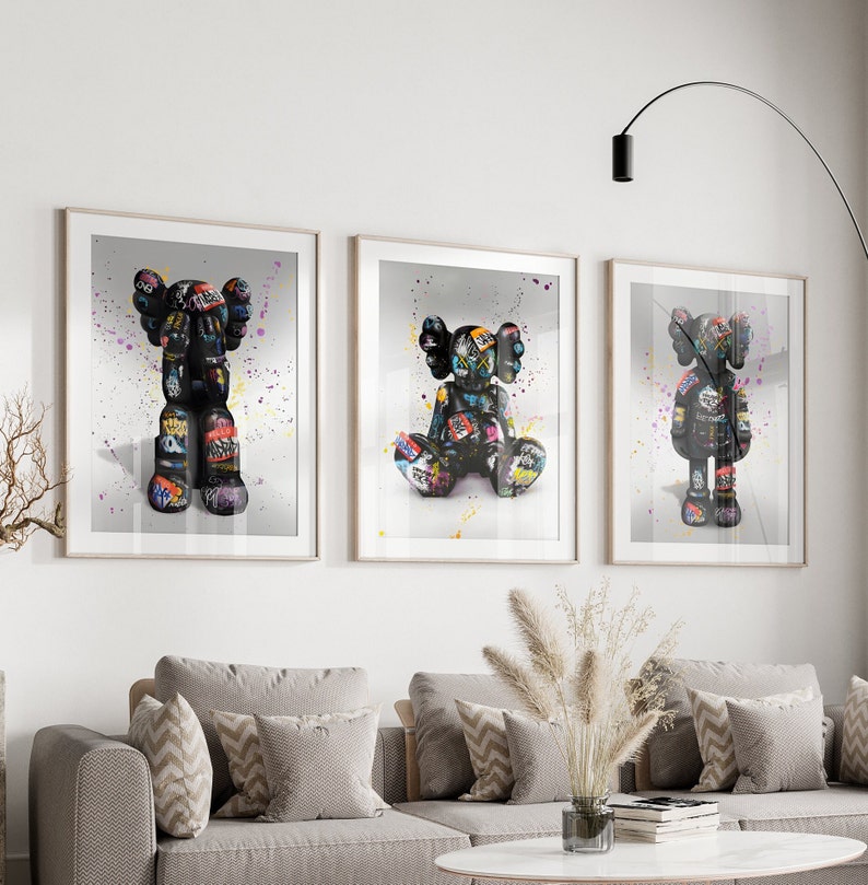 May include: Three framed prints of black and white graffiti art featuring a cartoon character. The character is depicted in three different poses: standing, sitting, and with arms raised. The background of each print is a light grey with colourful paint splatters.