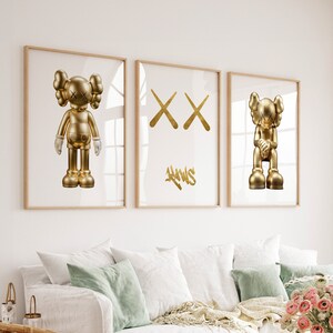 May include: Three framed prints featuring gold-coloured sculptures of a cartoon character. The character is depicted in three different poses: standing, with crossed arms, and sitting. The prints are hung on a white wall above a sofa.