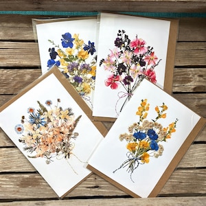 May include: Four greeting cards with dried flower bouquets. Each card features a different arrangement of colorful flowers, including blue, yellow, pink, and purple blooms, tied with twine. The cards are displayed in a wooden crate.