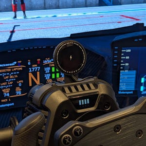 Op de afbeelding: Een close-up van een race-simulator cockpit, met een zwart en grijs bedieningspaneel en digitale displays. Een draadloze oplader met een bliksemsymbool is bovenop gemonteerd. De displays tonen racegegevens.