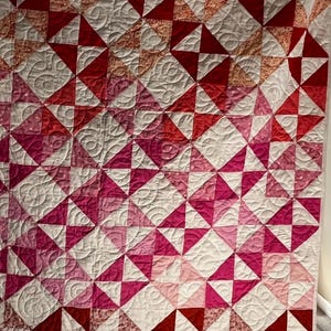 May include: A handmade quilt featuring a geometric pattern of red, pink, and white triangles. The quilt has a quilted design with a variety of shades of red and pink, creating a gradient effect. The quilt is a cozy and decorative home textile.