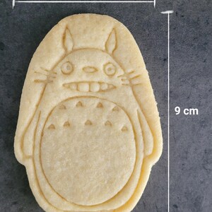 May include: A light-colored cookie in the shape of Totoro, measuring approximately 3.5 inches wide and 3.5 inches tall. The cookie features detailed facial and body features, including a smiling mouth and a dotted belly. The word "Totoro" is printed at the bottom.