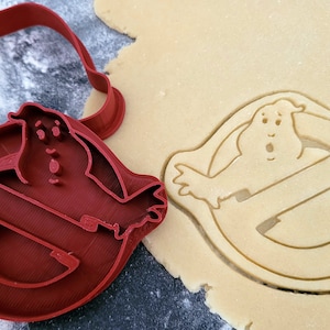 Puede incluir: Cortador de galletas de plástico rojo con forma del logotipo de Ghostbusters con un fantasma en su interior. El cortador está sobre una hoja de masa para galletas sin hornear con el logotipo cortado.