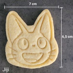 May include: A light-colored cookie in the shape of a cat's face, measuring 2.75 inches wide and 2.56 inches tall. The cookie has a smiling face with large eyes and the word "Jiji" at the bottom. The cookie is a baked good.
