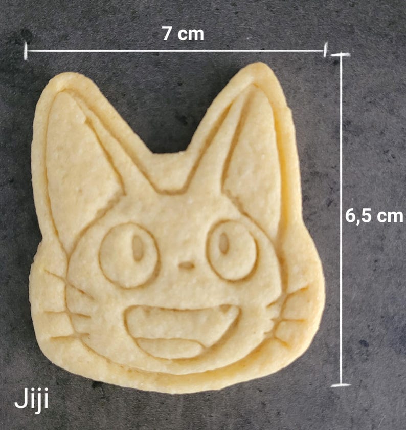 May include: A light-colored cookie in the shape of a cat's face, measuring 2.75 inches wide and 2.56 inches tall. The cookie has a smiling face with large eyes and the word "Jiji" at the bottom. The cookie is a baked good.