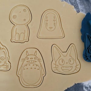 May include: Cookie cutters in various shapes pressed into dough. Designs include a forest spirit, a character with a blank face, a fire demon, Totoro, and a cat. A blue cat-shaped cutter is also present.