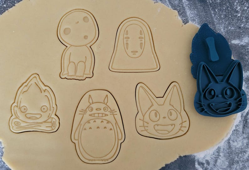 May include: Cookie cutters in various shapes pressed into dough. Designs include a forest spirit, a character with a blank face, a fire demon, Totoro, and a cat. A blue cat-shaped cutter is also present.