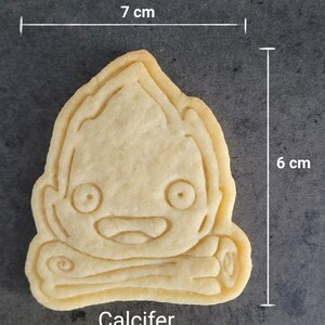 May include: A light-colored cookie in the shape of a cartoon character, measuring 7 cm wide and 6 cm tall. The cookie has a smiling face and the word "Calcifer" at the bottom.