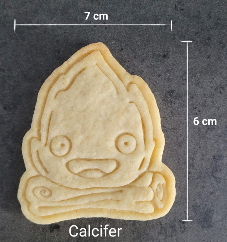 May include: A light-colored cookie in the shape of a cartoon character, measuring 7 cm wide and 6 cm tall. The cookie has a smiling face and the word "Calcifer" at the bottom.