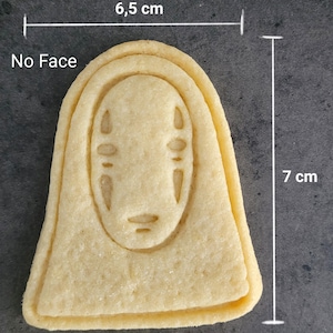 May include: A light-colored cookie shaped like the character "No Face" from the animated film. The cookie is 7 cm tall and 6.5 cm wide. The face details are subtly imprinted. The cookie is on a dark gray surface.