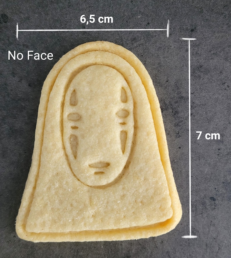 May include: A light-colored cookie shaped like the character "No Face" from the animated film. The cookie is 7 cm tall and 6.5 cm wide. The face details are subtly imprinted. The cookie is on a dark gray surface.