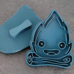May include: A teal-colored, 3D-printed cookie cutter set featuring a cartoon campfire design. The set includes the campfire-shaped cutter and a matching handle for easy use. The campfire has a smiling face and flame details.