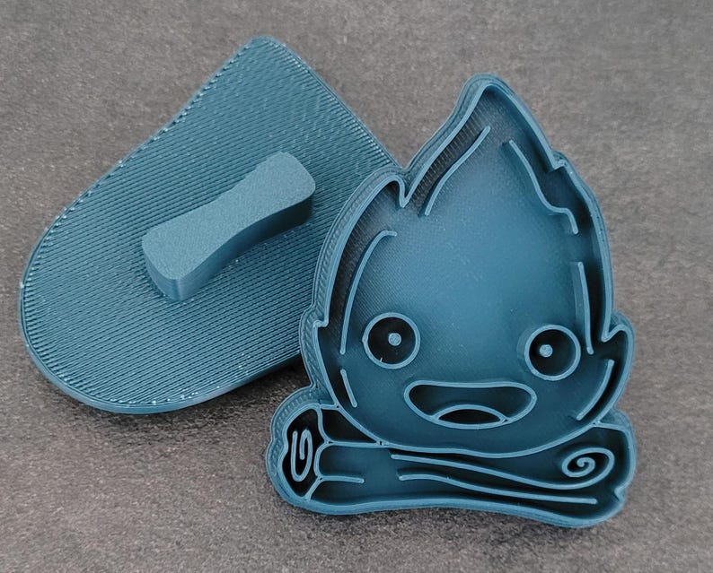 May include: A teal-colored, 3D-printed cookie cutter set featuring a cartoon campfire design. The set includes the campfire-shaped cutter and a matching handle for easy use. The campfire has a smiling face and flame details.